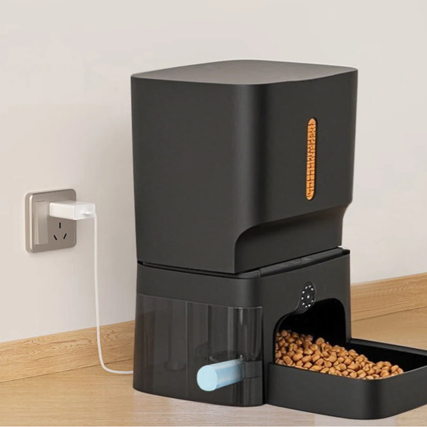 Black pet feeder with food on a wooden surface, plugged into an outlet.